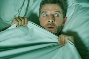 Man in bed with the sheet pulled up to his chin looking scared.